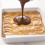 Melted peanut butter chocolate topping being poured over the no bake peanut butter bar base.