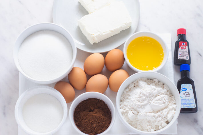 Bowls of ingredients for red velvet brownies including eggs, flour, Dutch-process cocoa powder, cream cheese, and red food coloring.
