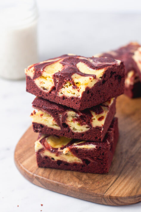 A high vertical stack of three Red Velvet Cheesecake Brownies on a wooden board, showcasing the thick, fudgy red velvet base and the swirled cream cheese topping.